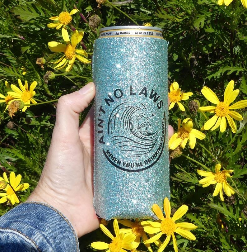 A hand holding the blue glitter koozie that features the White Claw logo and the words "Ain't no laws when you're drinking the Claws" around it