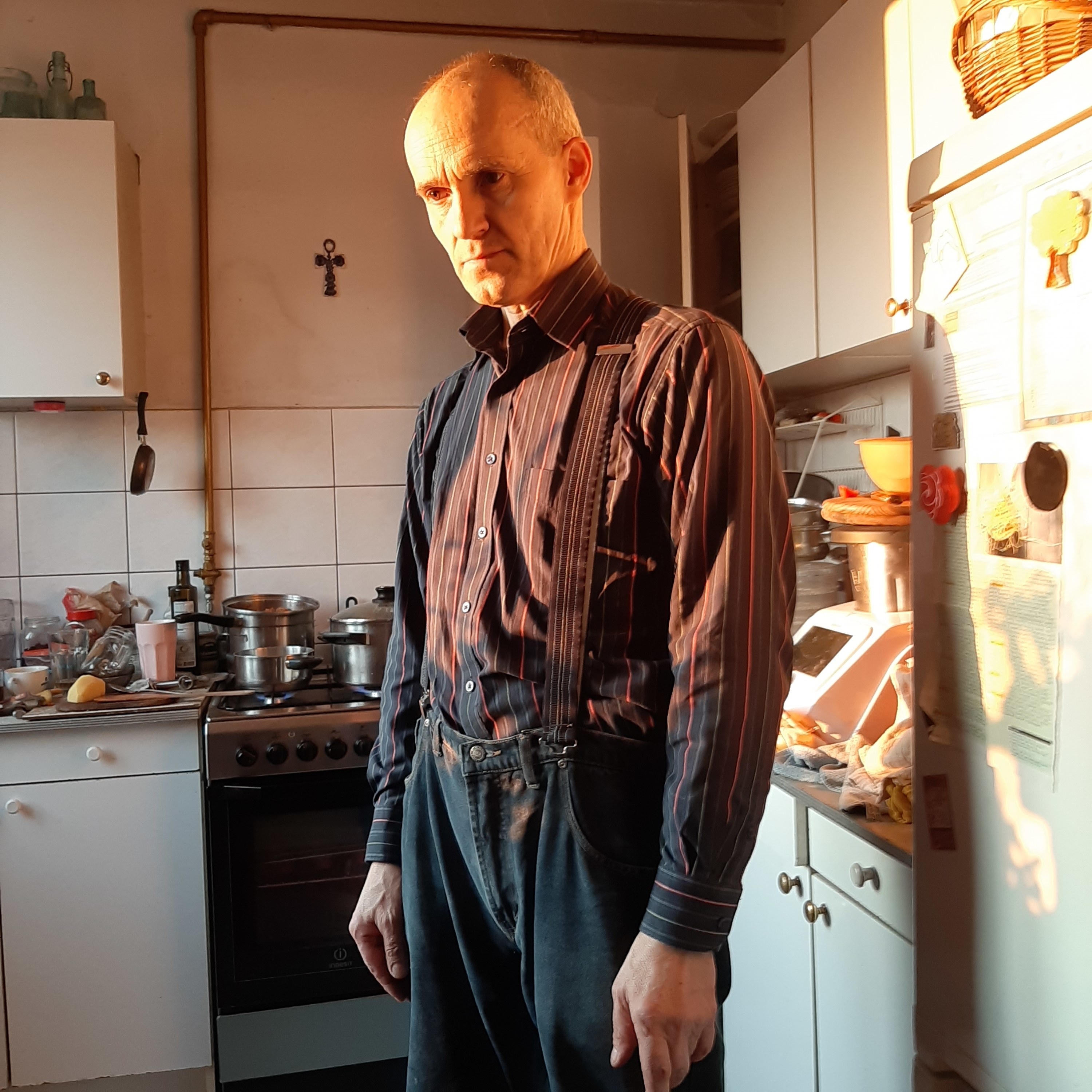 A man in overalls and a striped shirt stands in a kitchen