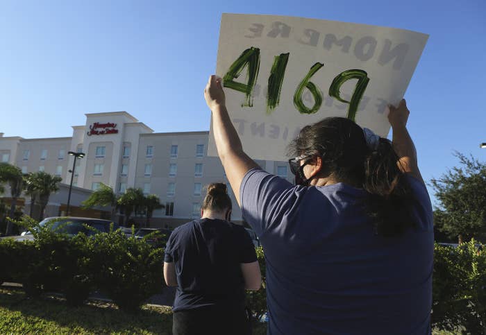 A person with their back to the camera holds up a protest sign, facing a Hampton Inn hotel