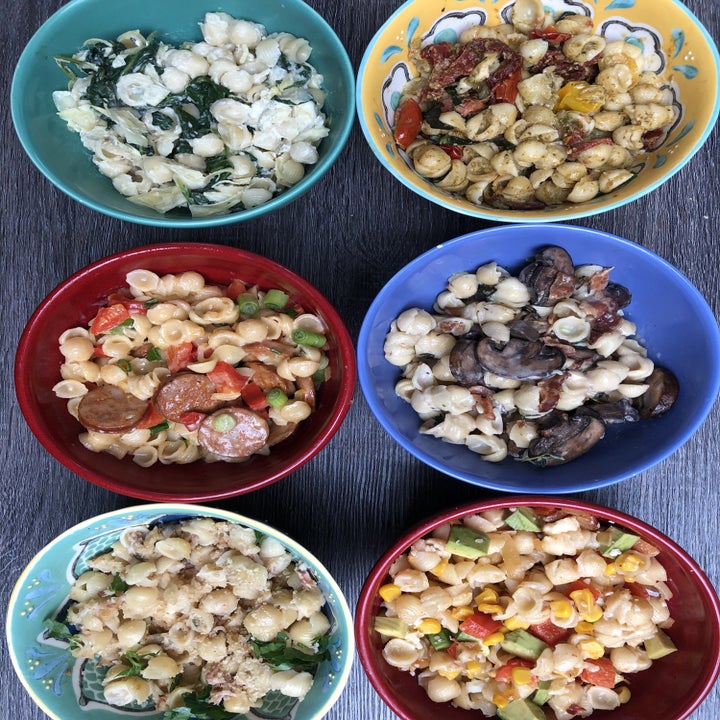 Six colorful bowls of doctored up mac 'n' cheese with different ingredients.