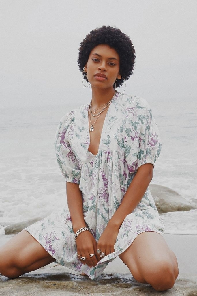 A model kneeling in ocean waves with the dress billowing in the breeze and the large, rounded sleeves hitting right at their elbow. The neckline plunges down to the sternum and the pattern is a purple and turquoise watercolor marble over the white base. 
