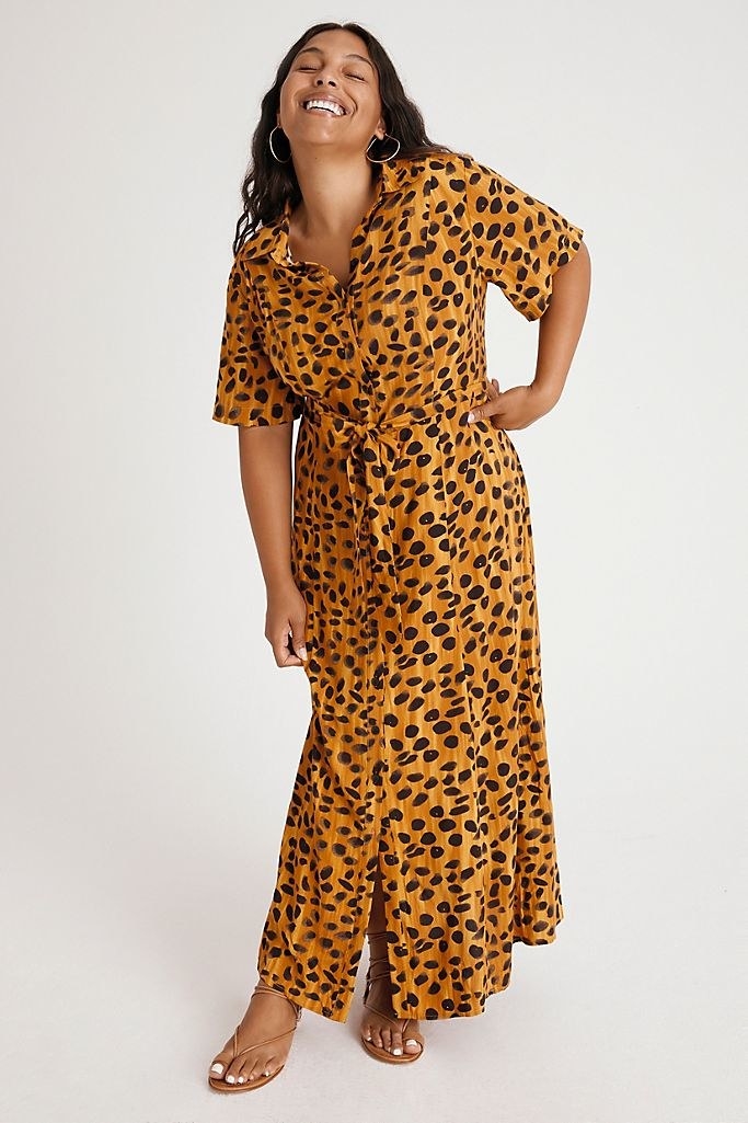 A model wearing the painted-animal-print dress with strappy sandals. There is a standard shirt collar and sleeves, with buttons down the front and a tie at the waist. 