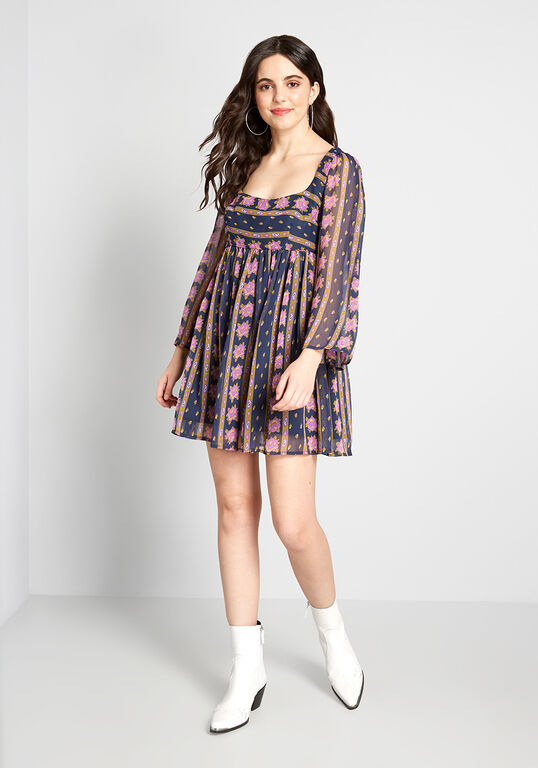 A model wearing the mini dress with ankle-high white boots. The dress is flowy with a striped floral pattern, a square neckline, and long sheer sleeves. 