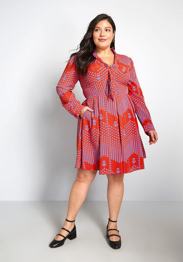 A model wearing the bright red and blue dress with black shoes. The dress has a geometric pattern, is knee length, has a fitted waist, a tie neckline, and long sleeves. 