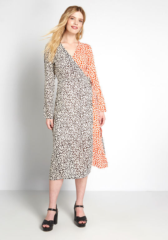A model wearing the dress with high heel sandals. The wrap dress ties at the side and has black animal print spots on the right half with orange spots on the left. 