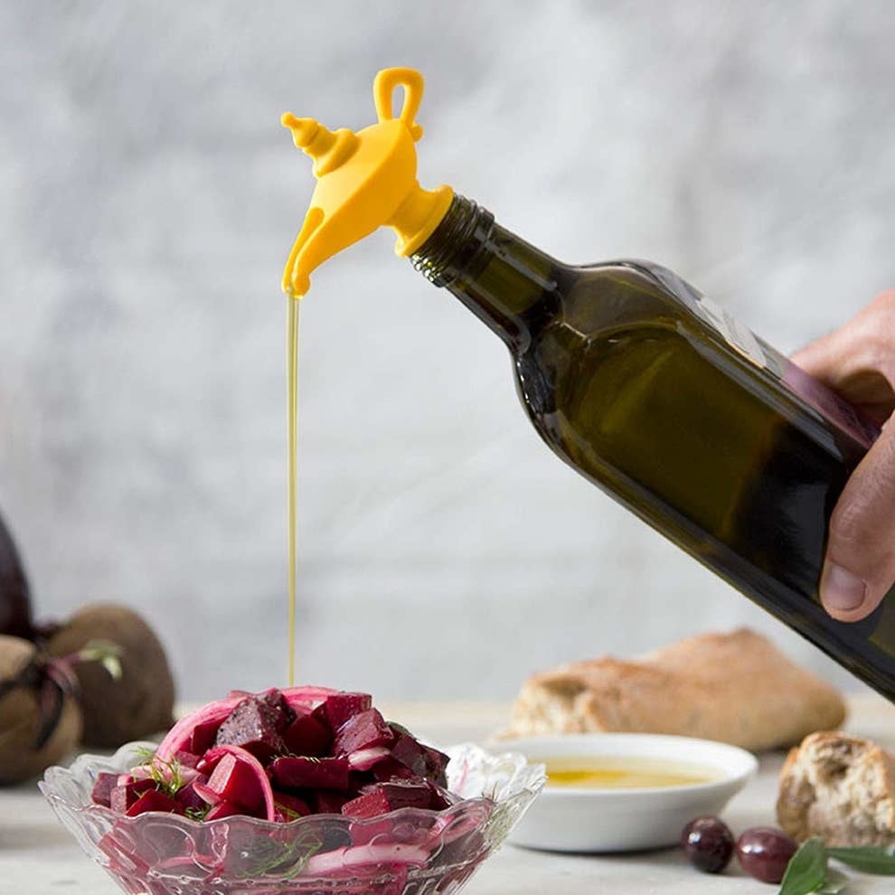 The stopped that looks like a genie lamp in yellow on top of a bottle of olive oil with some pouring out