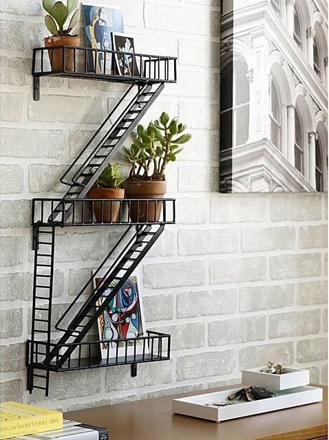 The fire escape shelf hanging above a desk, holding small potted plants and art prints