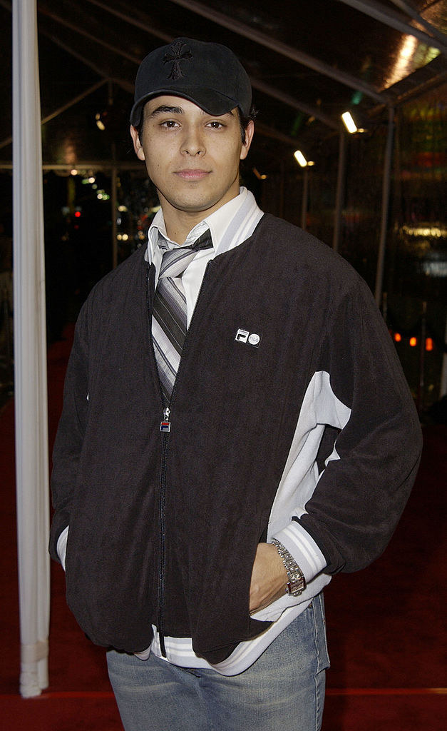 A photo of Wilmer Valderrama wearing a large striped tie worn with a jeans, an untucked button down, and velour jacket. 