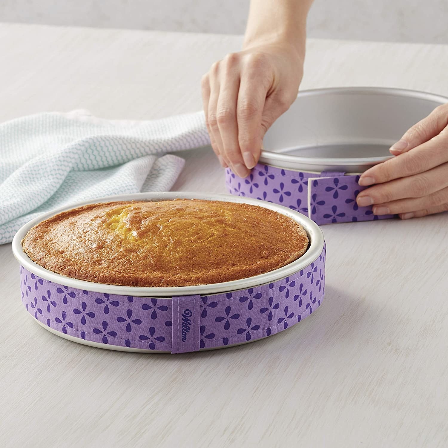 A person attaches the cake strips to a baking pan while another perfectly cooked cake rests next to it