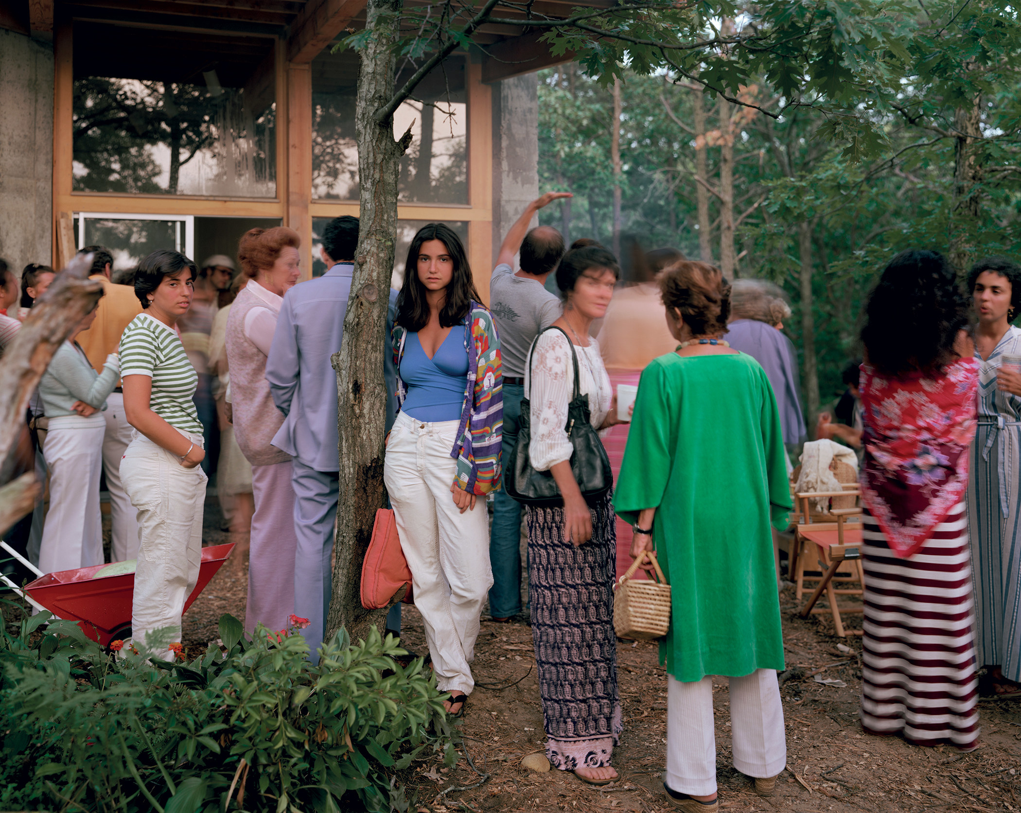 A young woman stands next to a tree and stares into the camera while surrounded by people at a crowded function