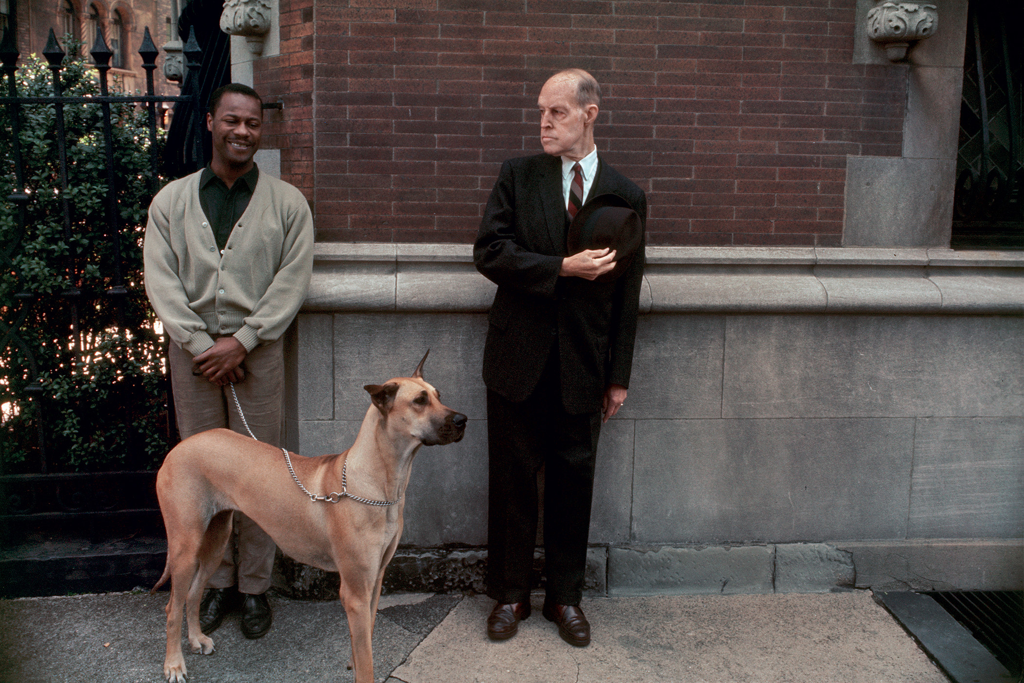 A man looks on at a second man holding a dog on a chain leash