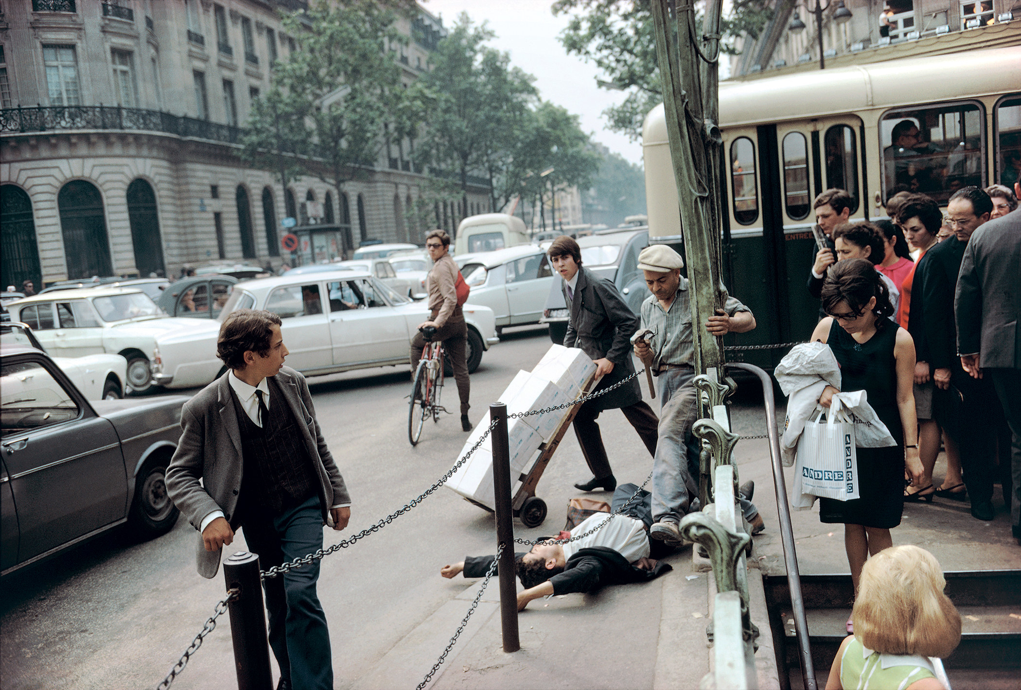 A man lies on the ground in the middle of a crowded city street while a cyclist and pedestrians look at him