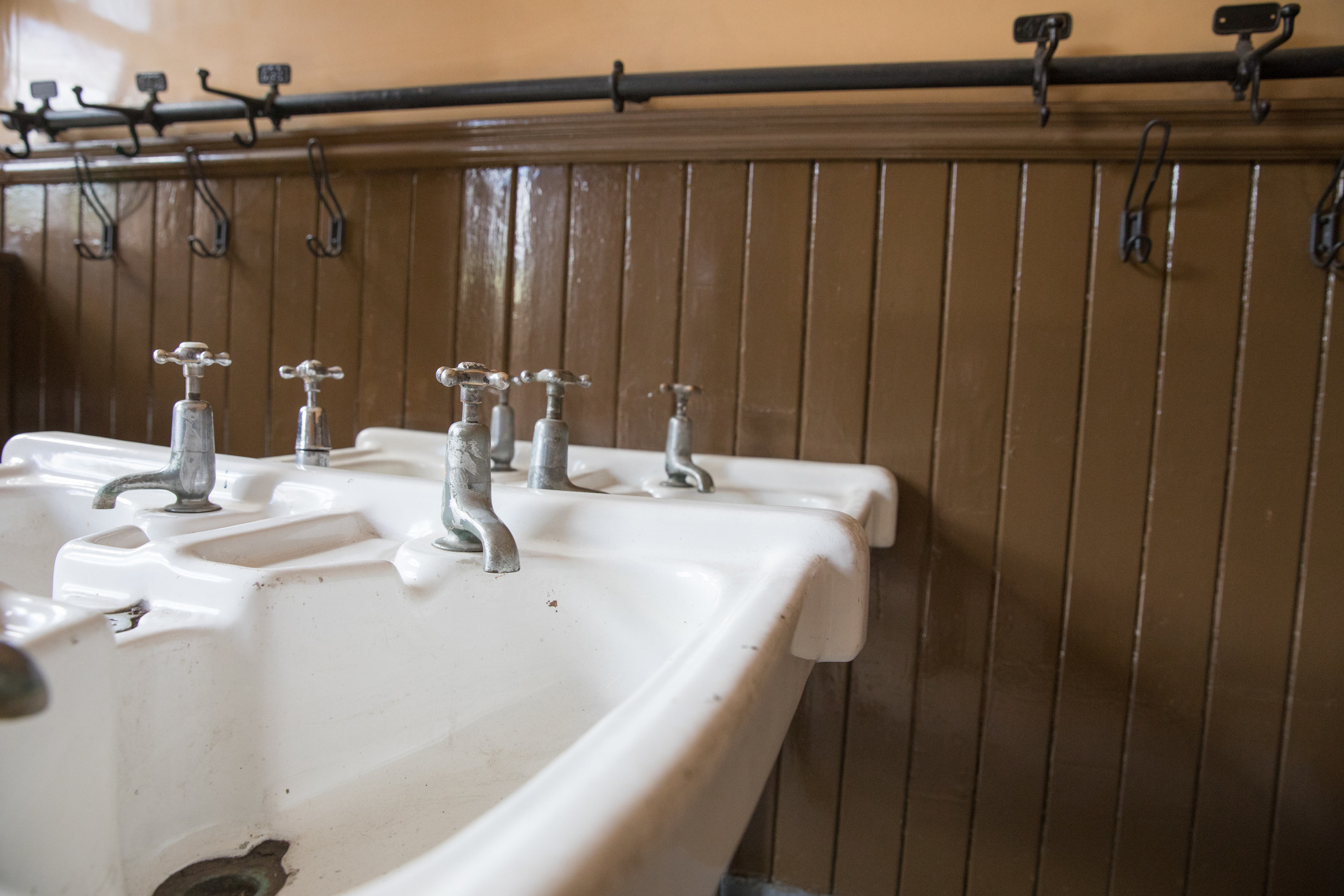 Old school sinks in a school washroom with pegs in the distance