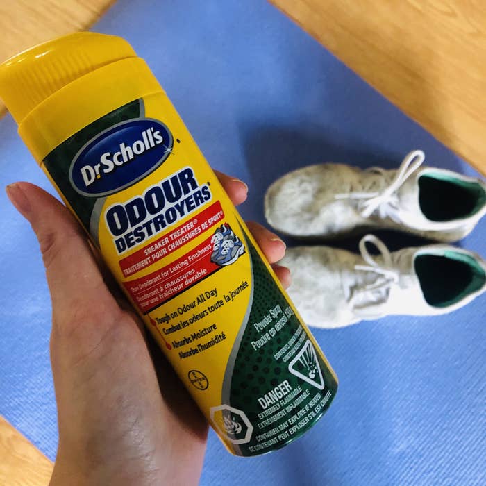A person holding a bottle of Dr. Scholl&#x27;s Odour Destroyers Sneaker Treater in front of a pair of running shoes