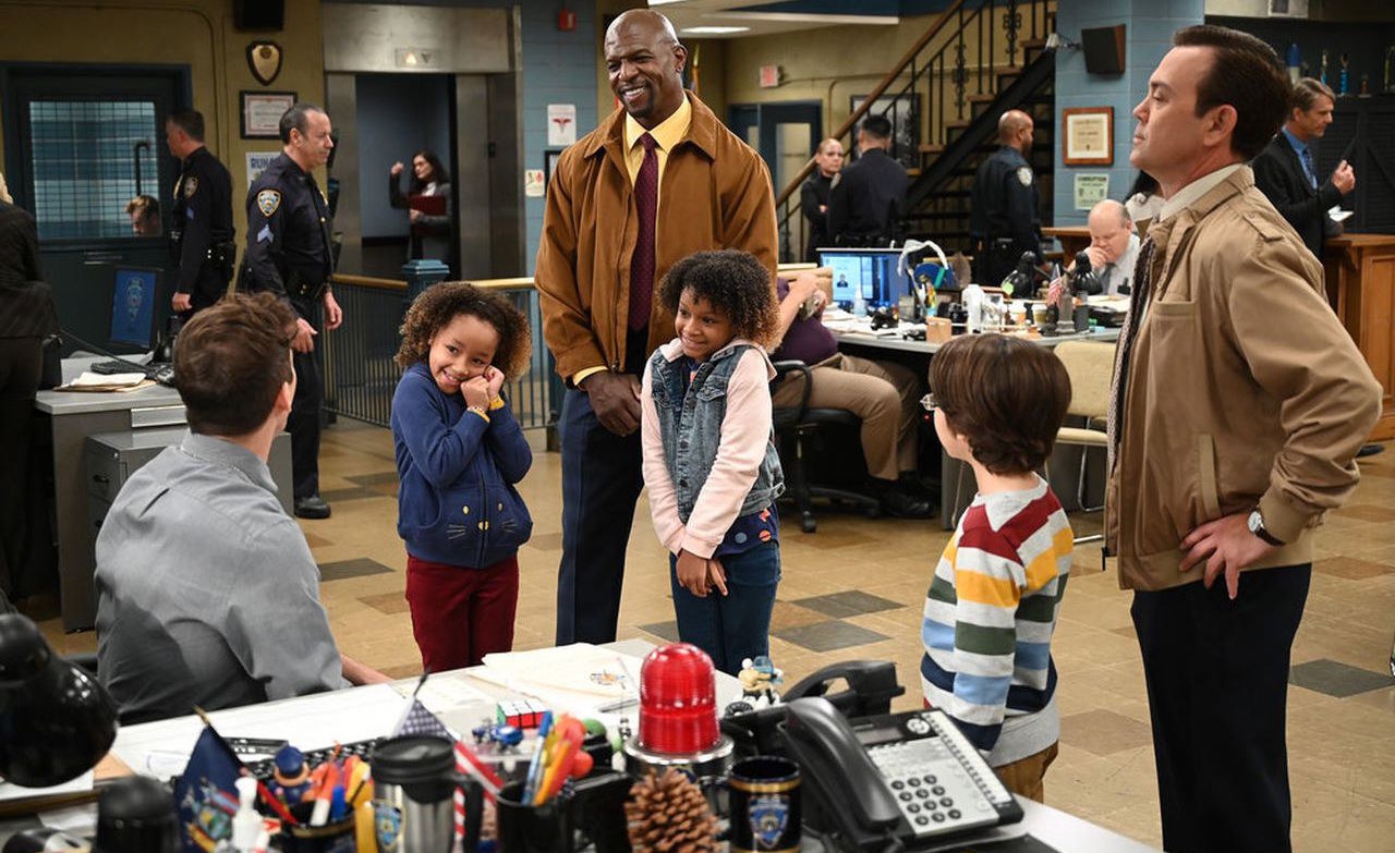 A still from Brooklyn 99 shows Jake, Terry, and Boyle with three children in their police precinct office area