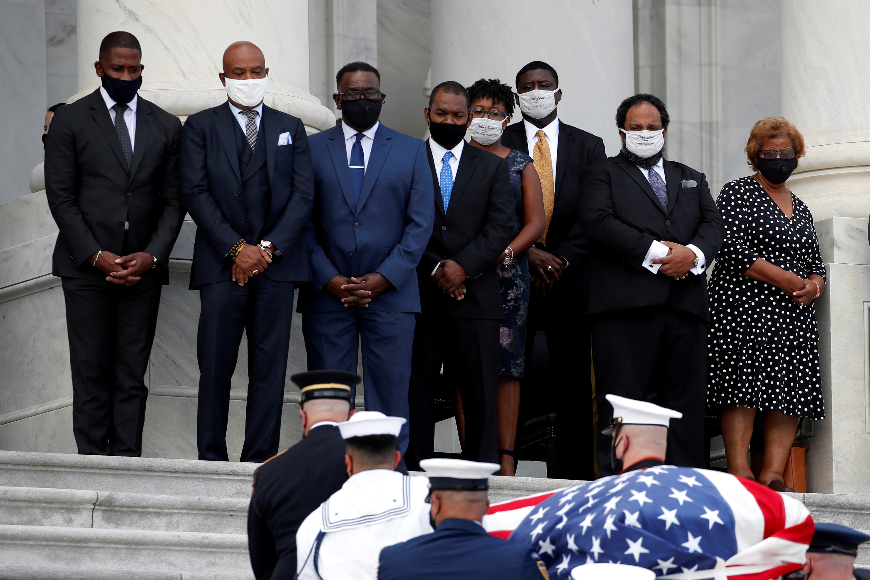 Several people wearing masks watch as a casket is carried by members of the military up a staircase