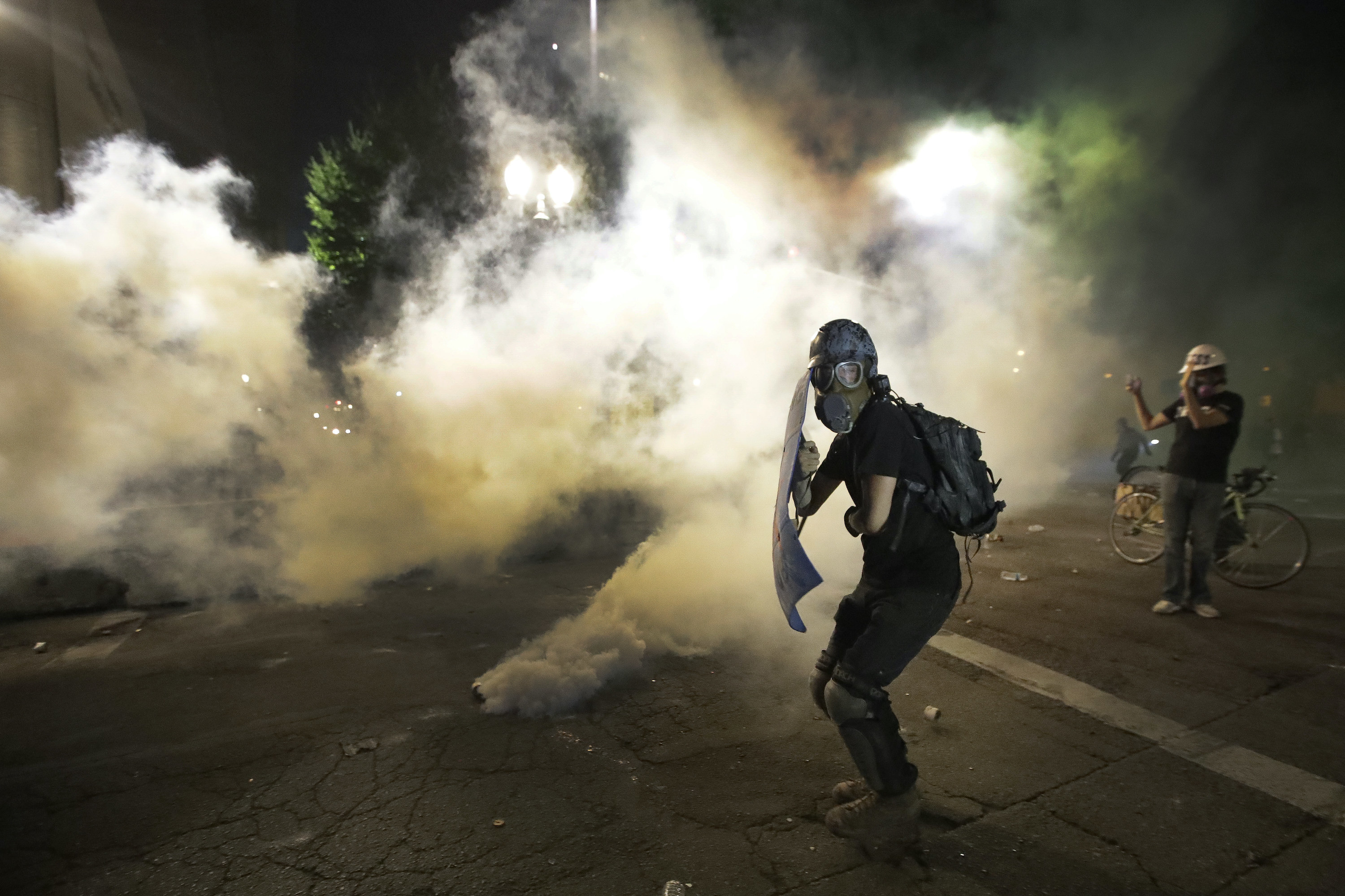 A protester holding a shield and wearing a gas mask hides from clouds of tear gas