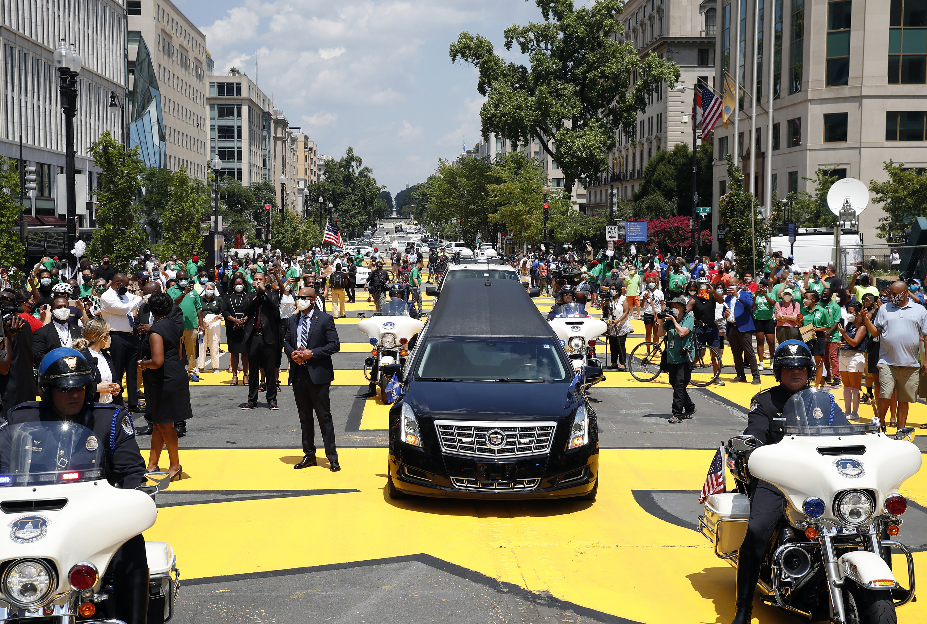 People spill into the street to surround a motorcade, including a hearse; big yellow letters on the street spell out &quot;Black Lives Matter&quot;