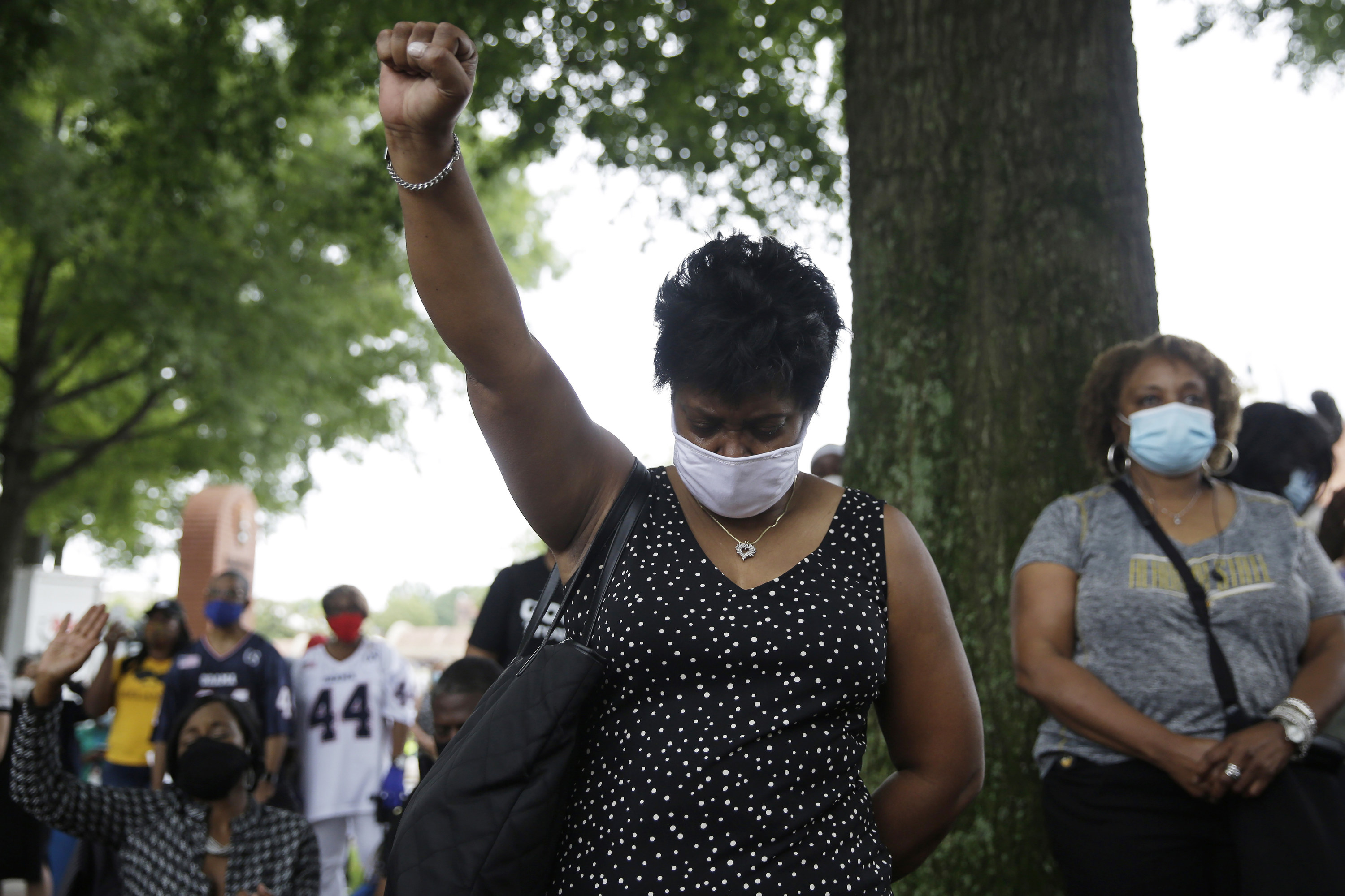 A black woman wearing a black dress and white mask looks to the ground and raises her fist