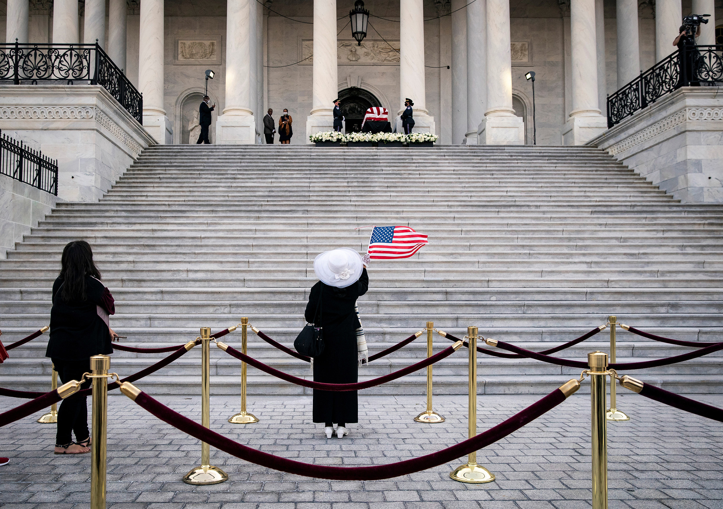 A woman dressed in black, with a white hat and white shoes, raises a US flag at the steps of the Capitol, looking up at John Lewis&#x27;s flag-draped casket at the top of the stairs