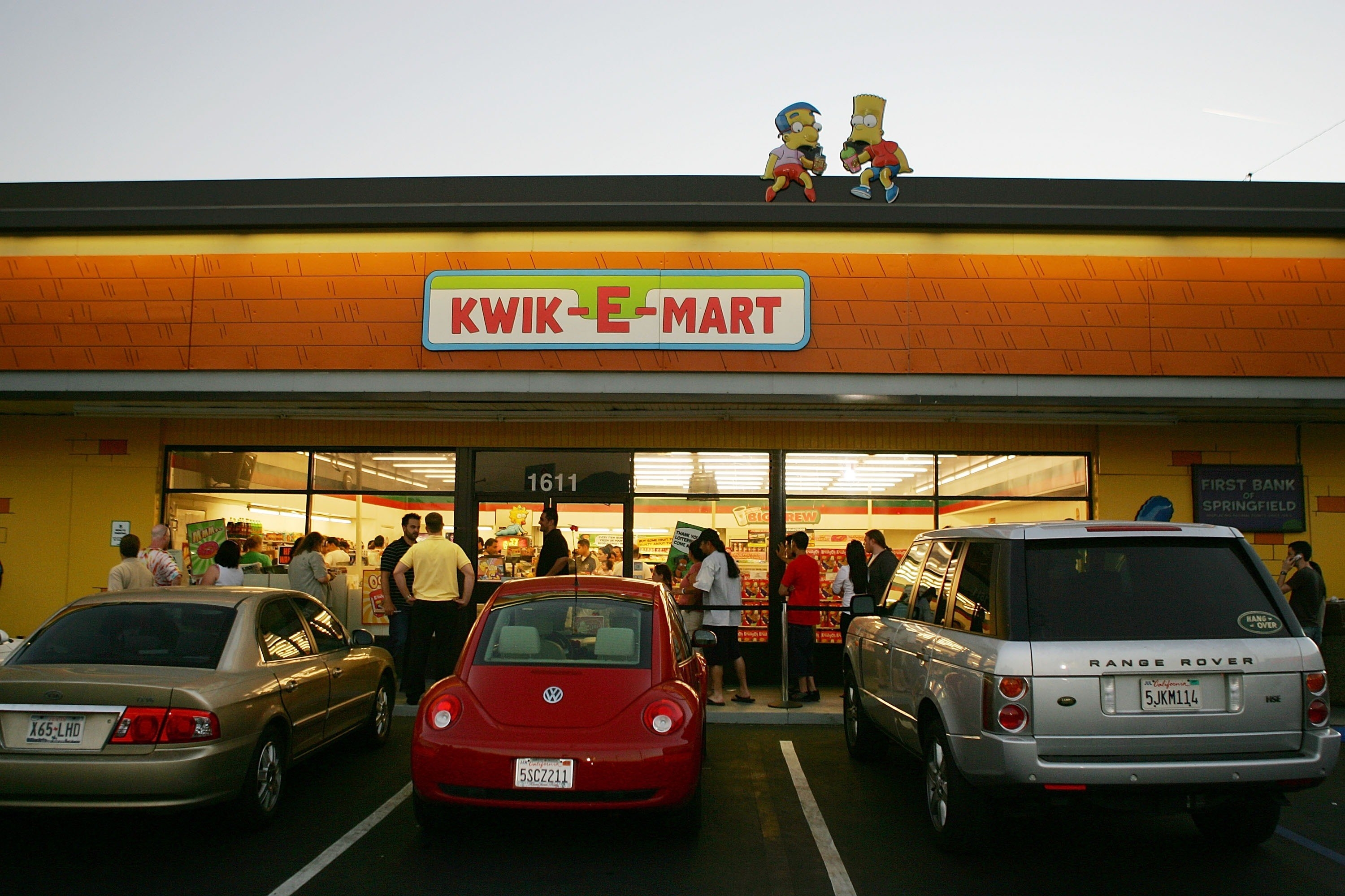 A exterior of a 7-Eleven store converted in a Kwik-E-Mart with a cardboard Milhouse and  Bart sitting on the roof.