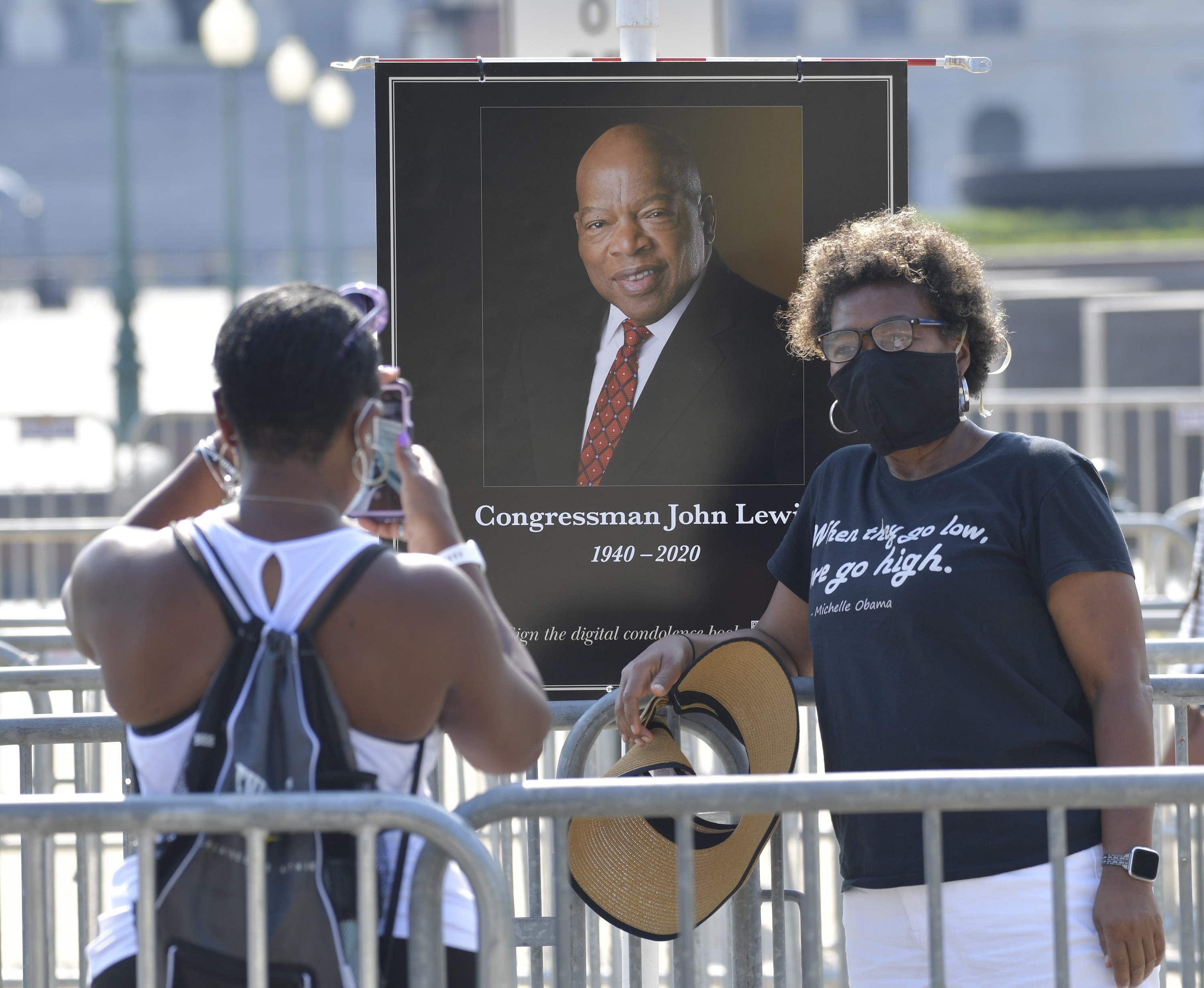 A woman takes a picture of another woman, who wears a mask and stands in front of a framed picture of John Lewis; her T-shirt reads a quote from Michelle Obama, &quot;when they go low, we go high&quot;