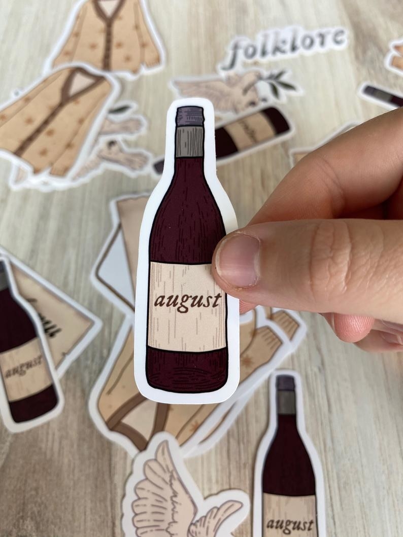 A hand holding a small sticker that&#x27;s shaped like a bottle of wine with a label that reads &quot;august&quot;
