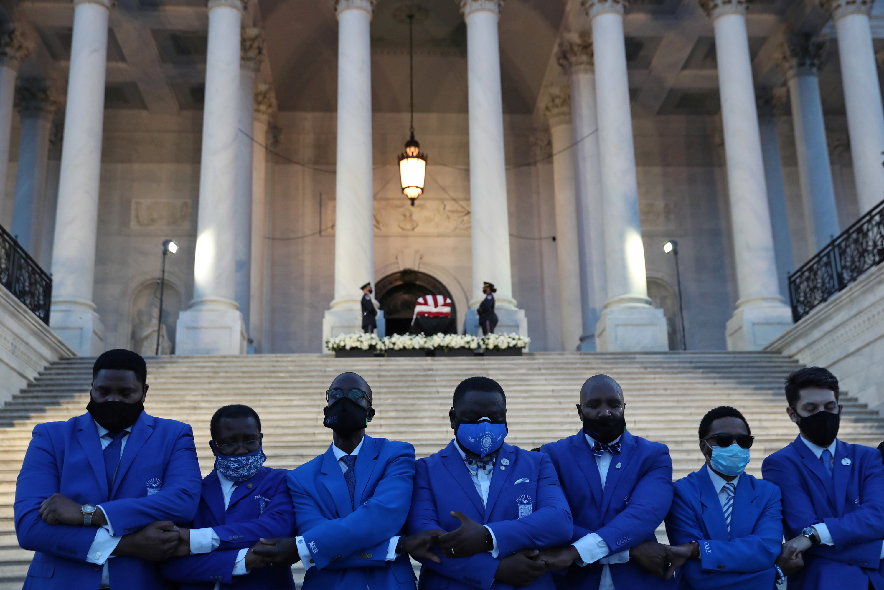 Men in blue suits, all wearing masks, stand shoulder to shoulder and hold hands outside the Capitol in Washington, DC