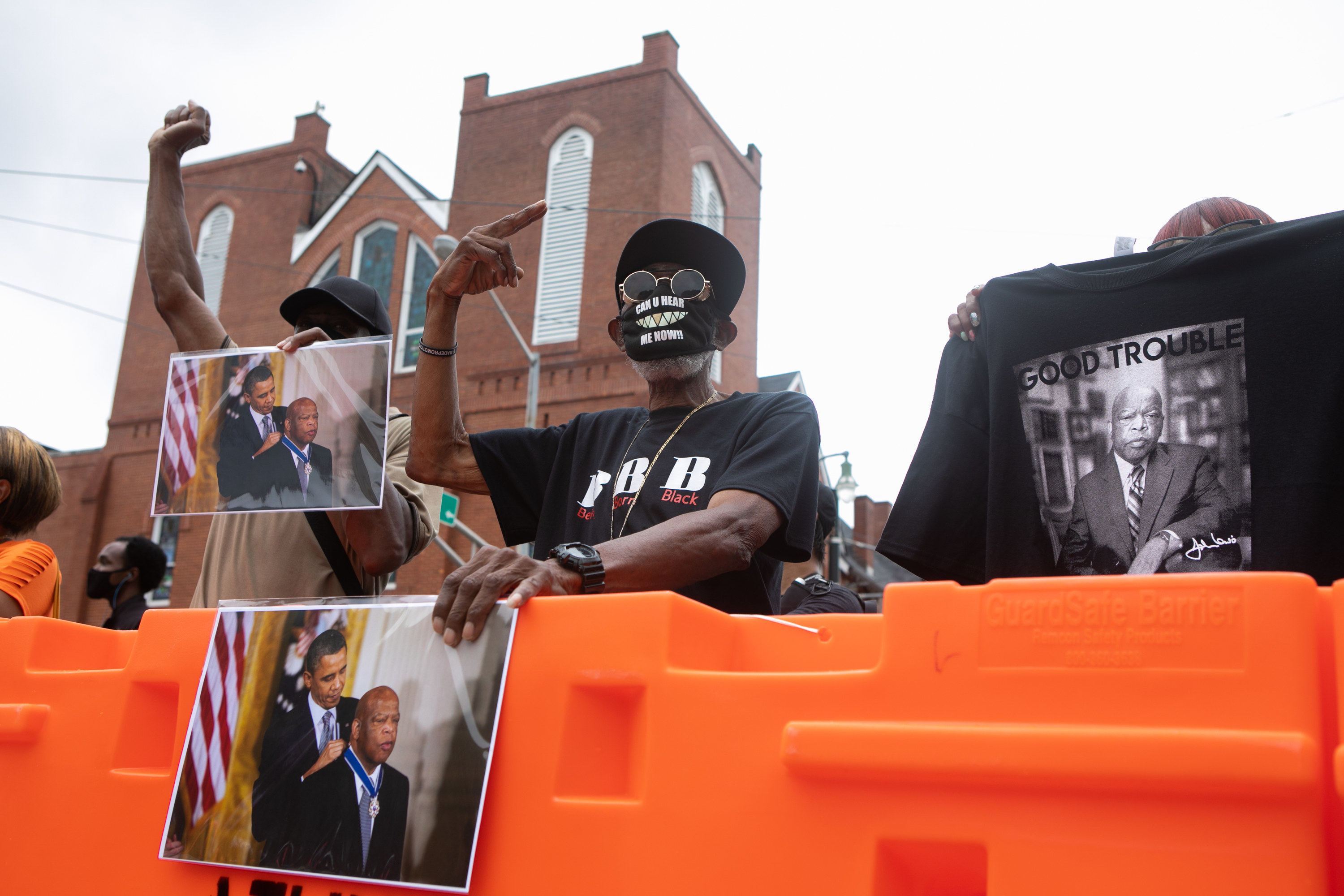 Two men hold images of former president Barack Obama giving John Lewis the Presidential Medal of Freedom; one of the men wears a mask that reads &quot;can you hear me now?&quot;