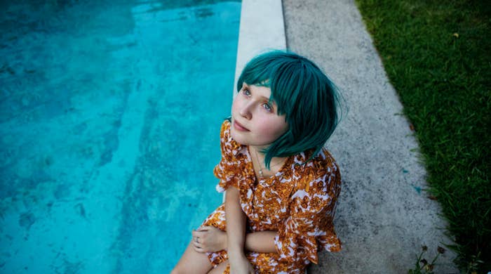 A girl in a short, bright wig sits by a pool, looking peacefully up at the sky