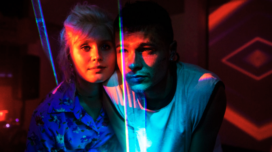 A young man and woman are close beside each other, technicolour lights projected onto them