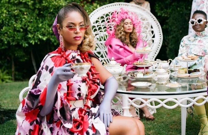 Beyoncé sits at a backyard table, which is filled with teapots and cookies for a tea party, and raises a teacup