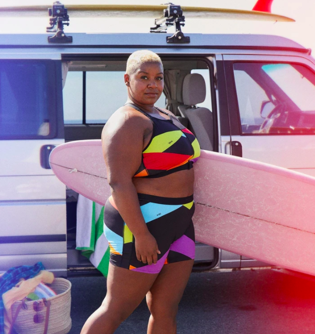 Model wears pair of neon color-block TomboyX board shorts while carrying a surfboard 