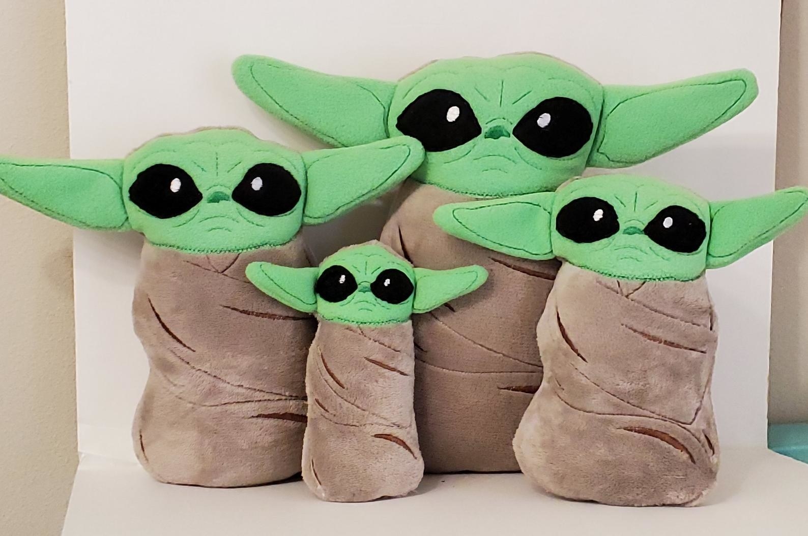 four different sized stuffed baby yoda dolls seemingly swaddled like a baby in a brown blanket