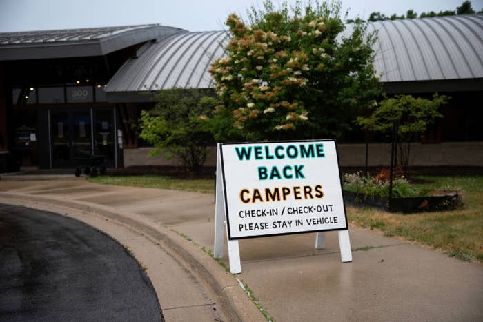 A sign on a sidewalk outside a building reads "Welcome back campers, check-in / check-out, please stay in vehicle"