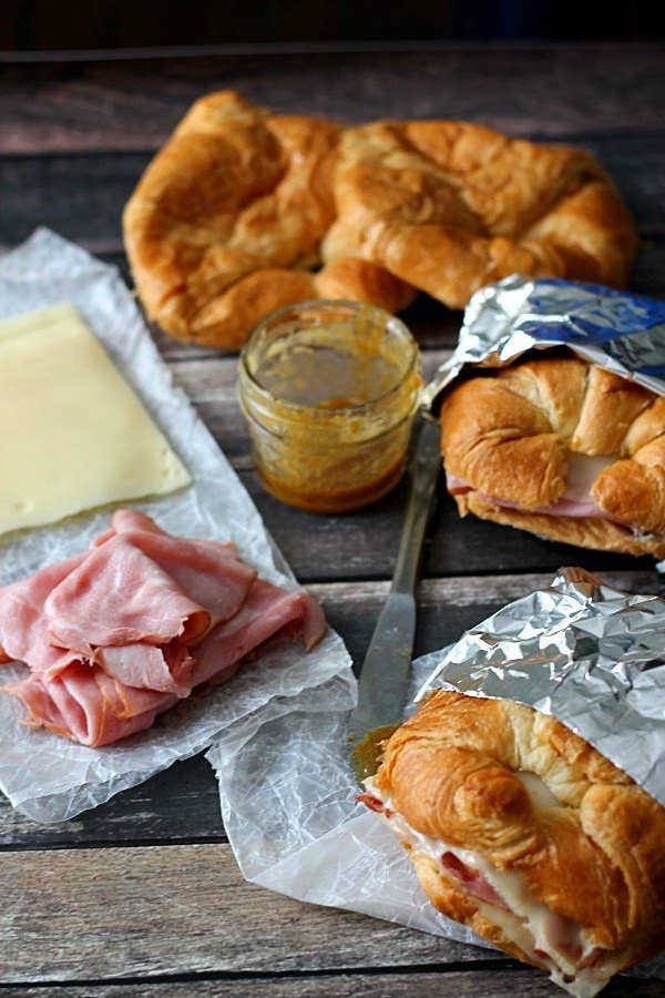 Ham, cheese, and honey mustard on a picnic bench, alongside two pre-made croissant sandwiches.