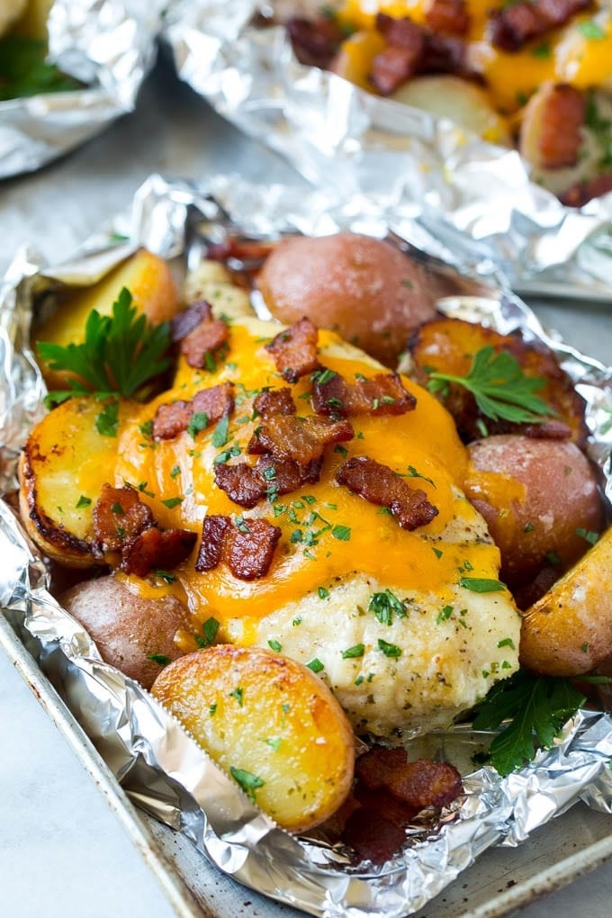A chicken breast covered in cheese and topped with bacon, surrounded by potato wedges in an aluminum foil pouch.