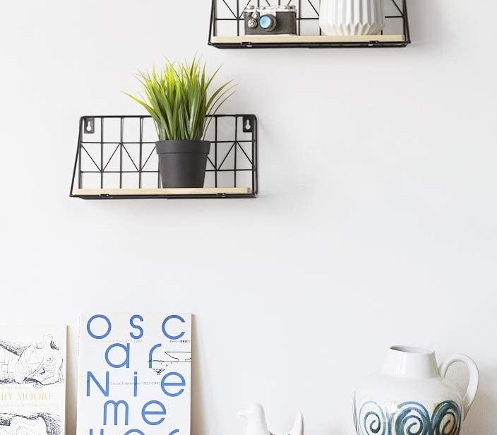 Two floating shelves on a wall with a wood base and a black wire back with assorted plants and trinkets on them