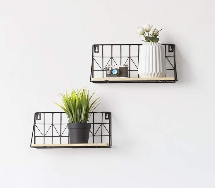 Two floating shelves on a wall with a wood base and a black wire back with assorted plants and trinkets on them