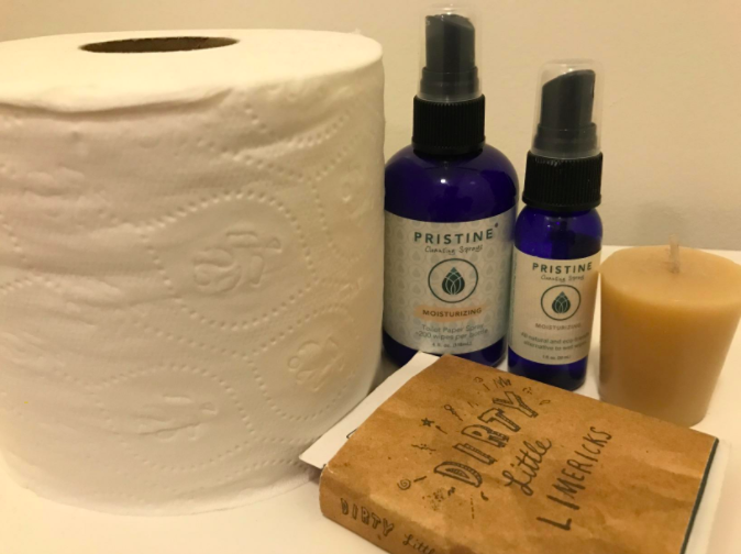 A large and small bottle of the product next to a roll of toilet paper, a book, and a candle in a bathroom