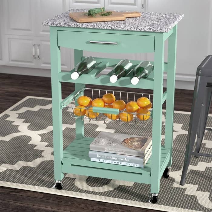A seafoam green kitchen cart with a shelf, bottle rack, metal container, drawer, and a gray granite top