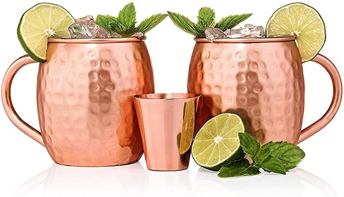 Two copper mugs are filled with liquid topped with ice mint and lime wedges with a copper shot glass