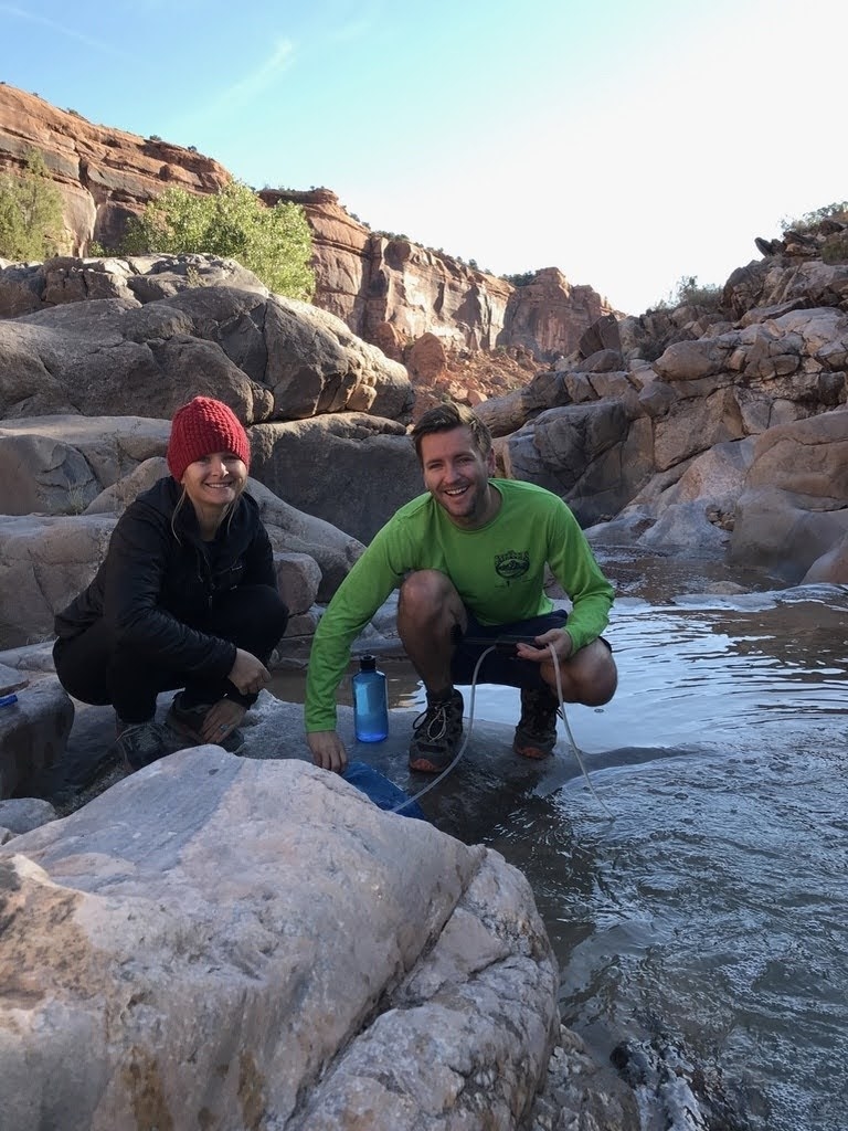 a couple filling up their water bottles from a flowing stream surrounded by rocky terrain