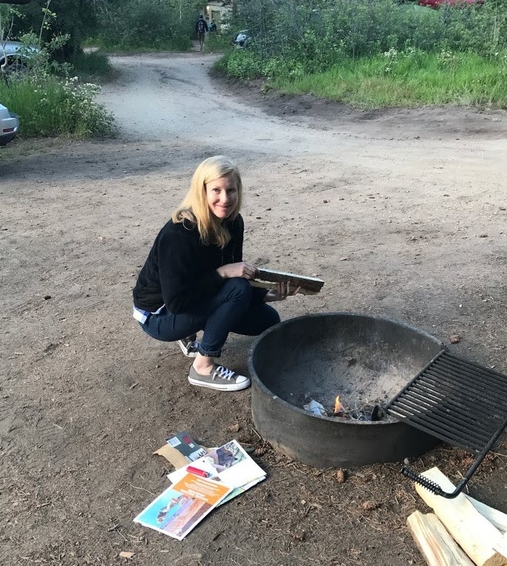 woman sitting by a metal campfire, with papers and firewood, getting ready to start a fire. 