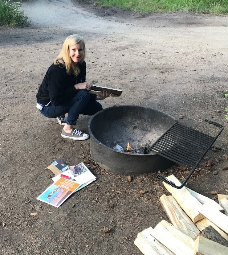 woman sitting by a metal campfire, with papers and firewood, getting ready to start a fire.
