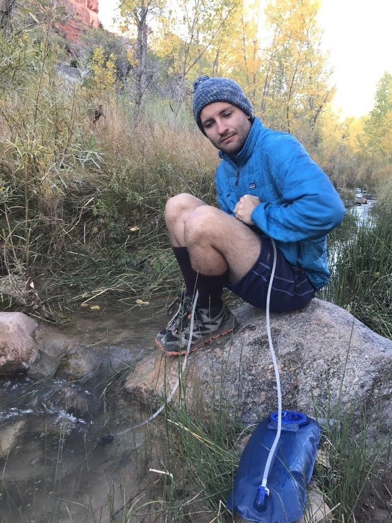 man sitting by a stream filling up his camel back bladder with water 