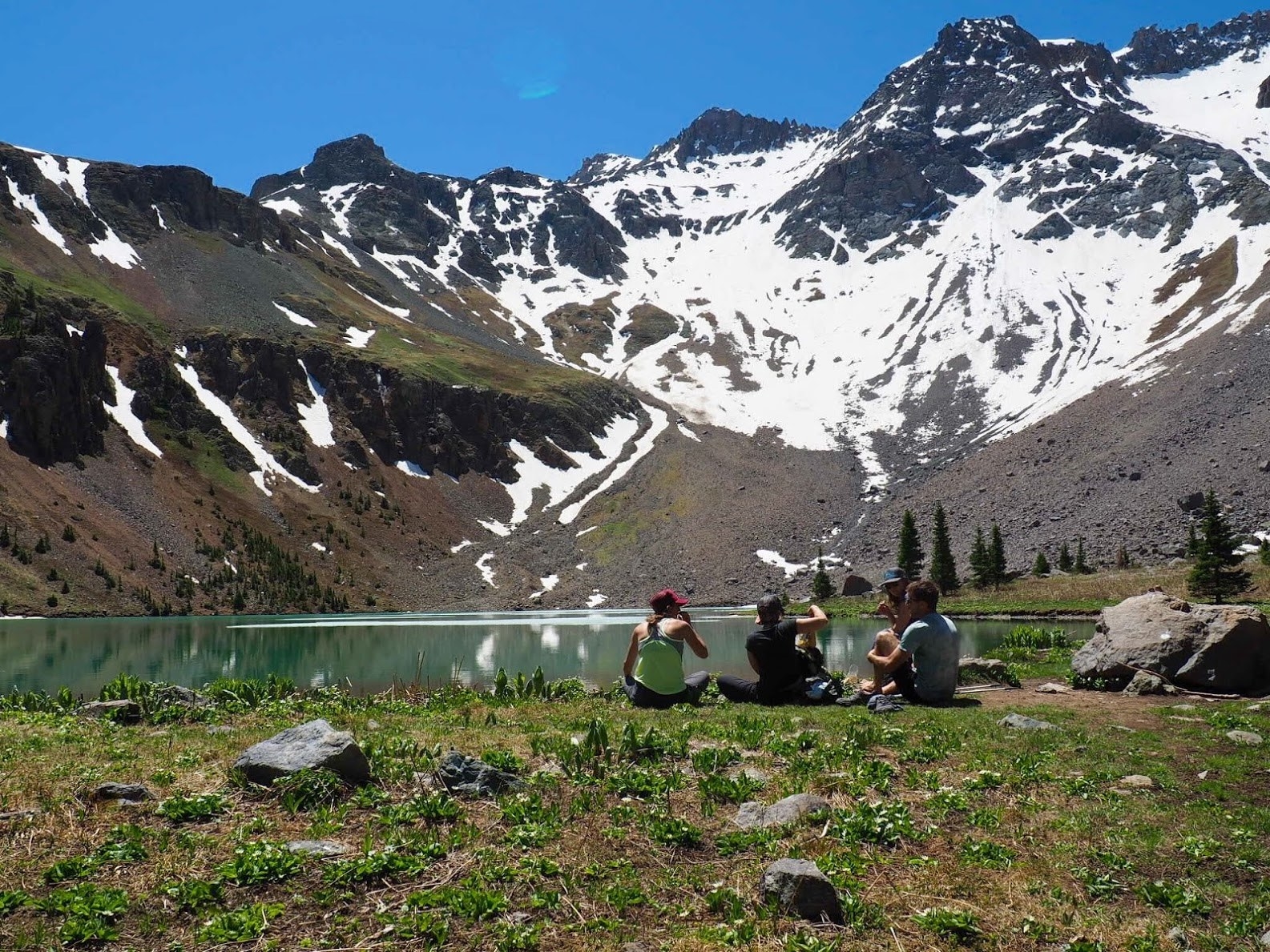 group of people sitting by a lake with a stunning snow-capped mountain in the background