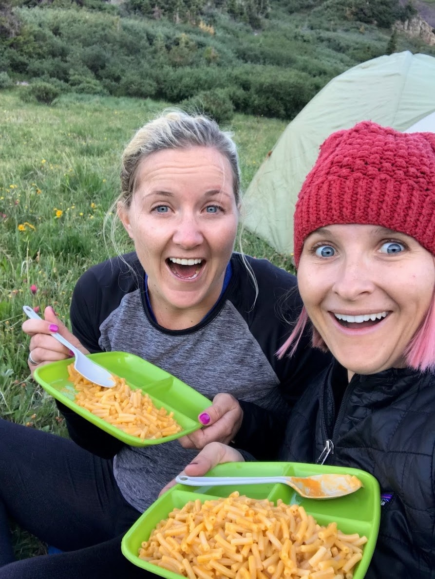 two women sitting on the grass eating packaged mac and cheese off plastic plates, smiling and taking a selfie