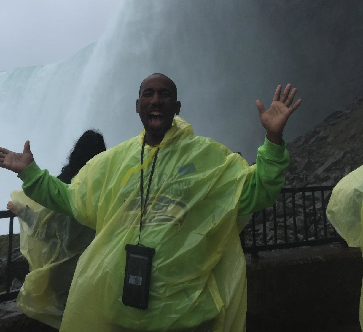 reviewer in poncho excitedly poses in front of niagara falls with phone in pouch around neck 