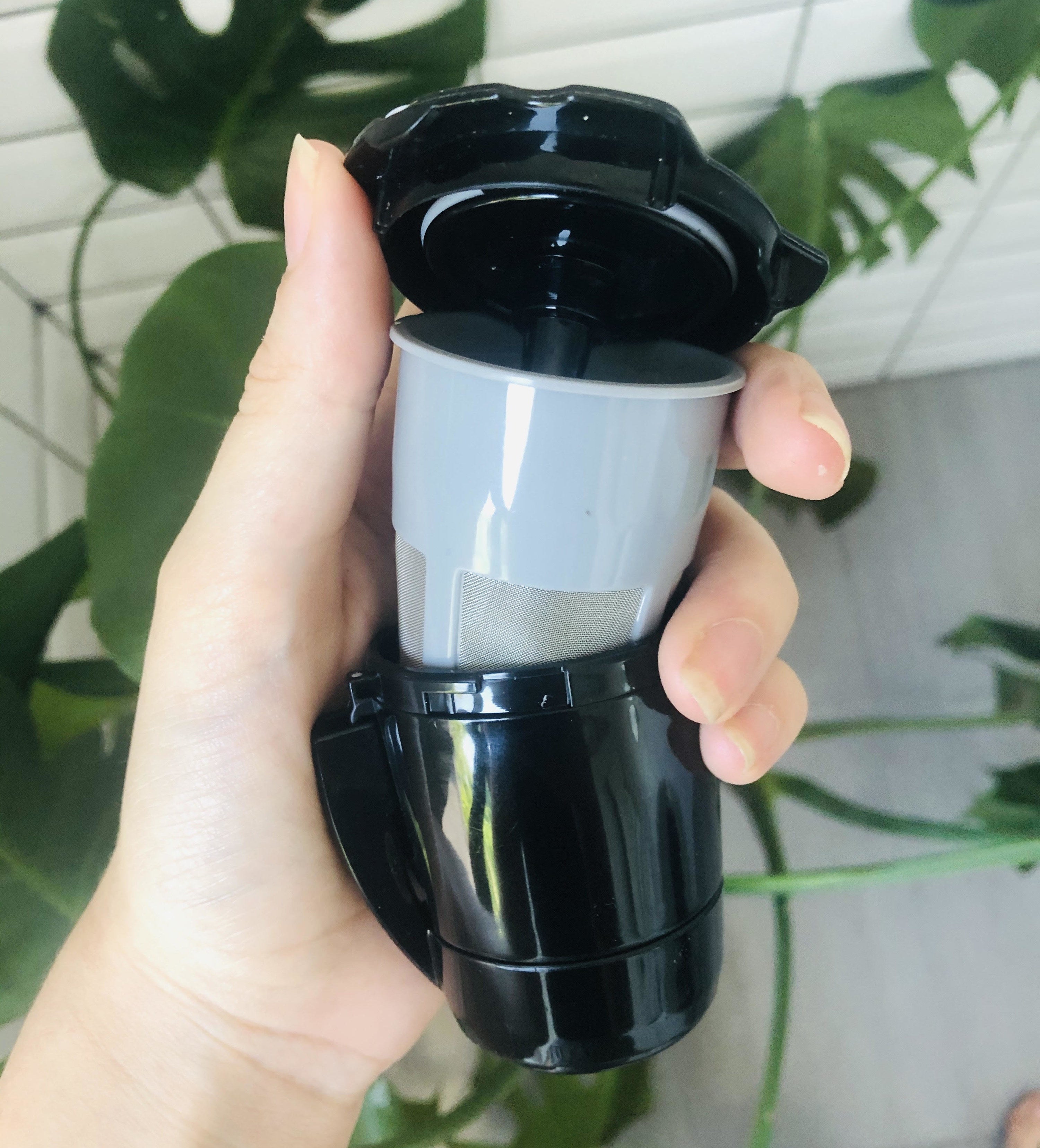 A person holding an opened reusable Keurig cup in front of a plant